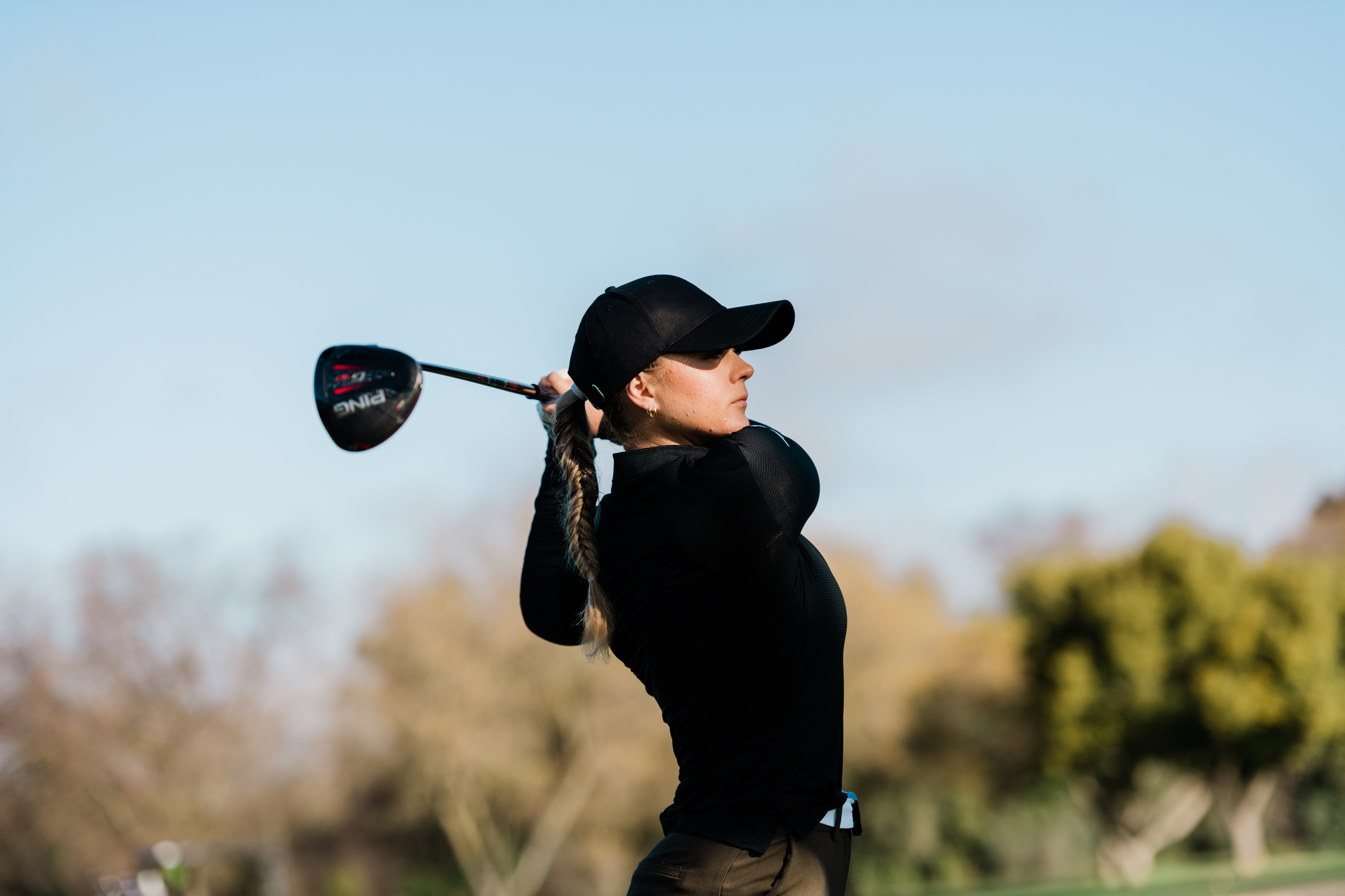 color image of a woman playing golf in black clothing and a black baseball hat, in mid swing, against a blue sky and yellow/green trees
