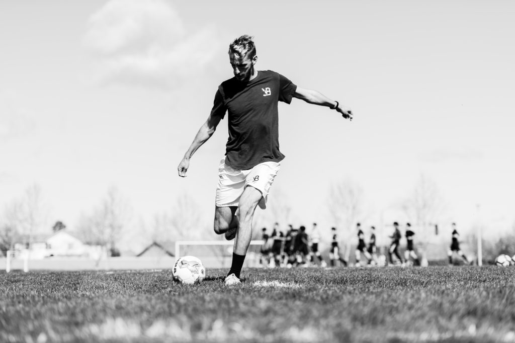 black and white image of a man in a soccer field in mid-stride, with arms outstretched, about to kick a soccer ball