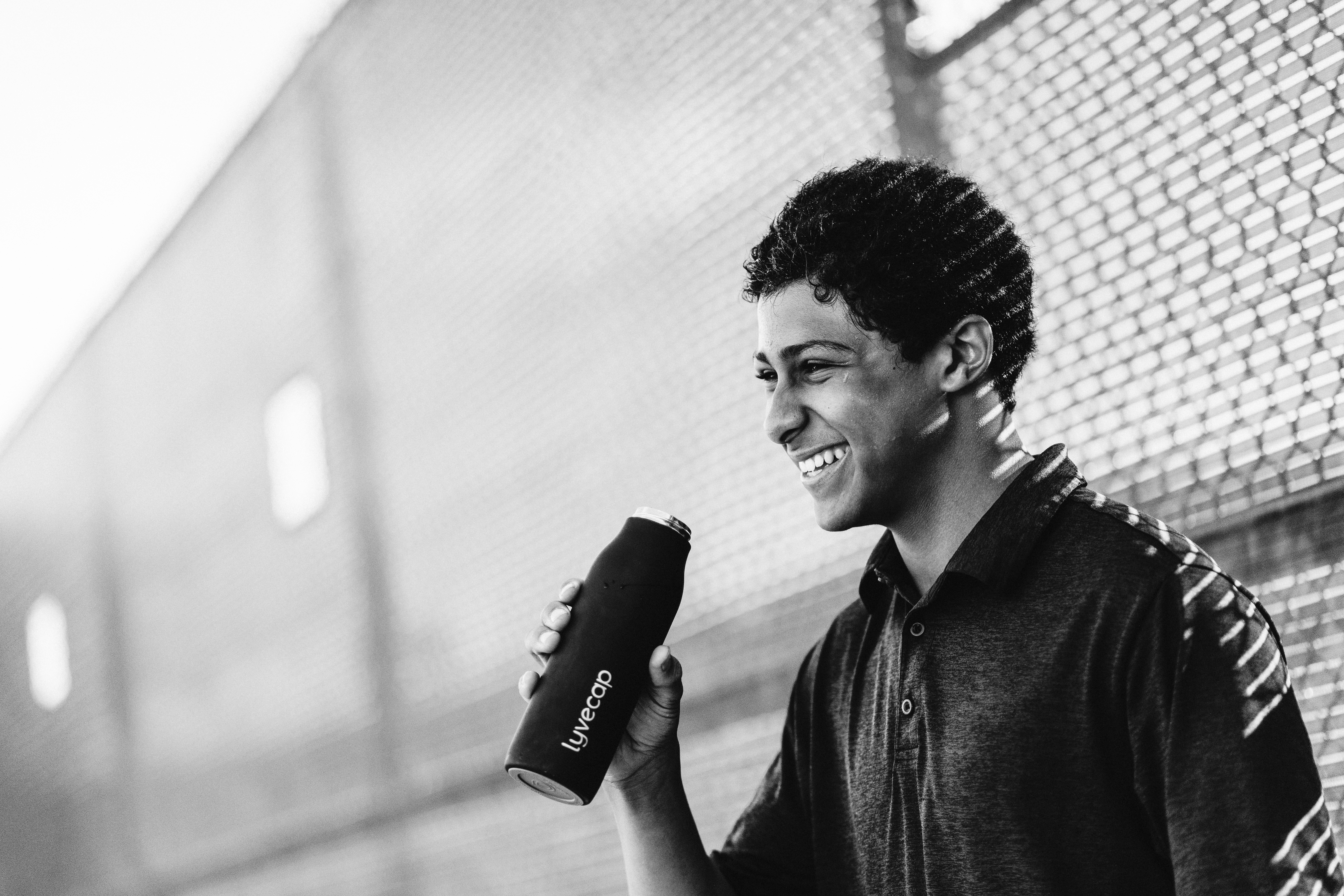 black and white image of a young man laughing against a chain link fence, while drinking out of a lyvecap probiotic bottle