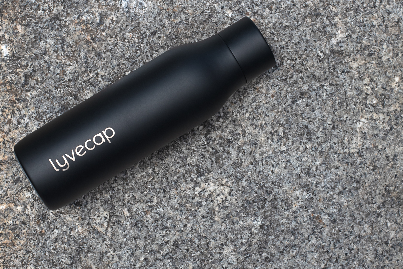 Image of a black Lyvecap bottle with a water drop logo laying on a grey speckled stone surface